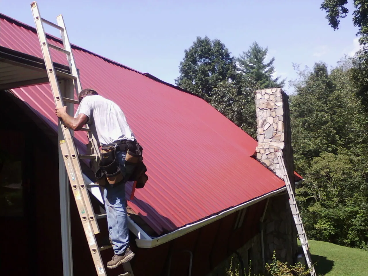 Roofer on a ladder beside a red metal roof and stone chimney — Soffit & Fascia Repair by Roofing Brothers in Saugus